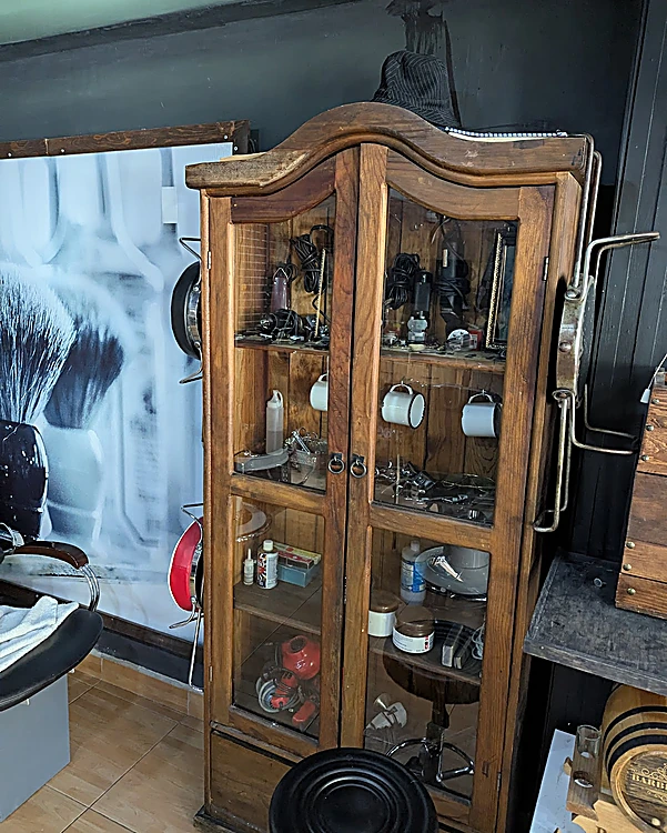 Vitrina antigua de madera con tazas, navajas y frascos; al fondo, la foto de una brocha de afeitar